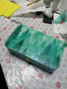 Na zdjęciu: Turkusowe, drewniane pudełko z wieczkiem ozdobionym obrazkiem choinek w odcieniach zielonego i turkusu.