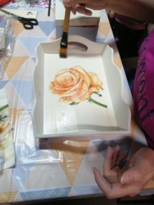 Na zdjęciu: prostokątna, biała trewniana taca z pomarańczowo - żółtą różą na dnie.