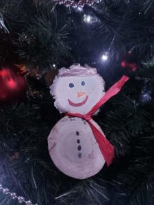 Na zdjęciu: Biały, uśmiechnięty bałwanek z czerwonym szalikiem. Wykonany z dwóch plastrów drzewa, zawieszony na choinkę.