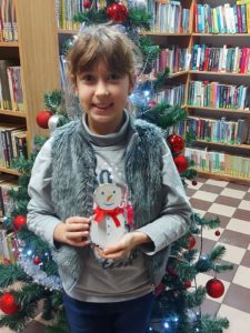 Na zdjęciu: Radosna dziewczynka w szarym serdaku. Trzyma w ręku bałwanka wykonanego z dwóch plasterków drzewa. W tle choinka i regały z książkami.
