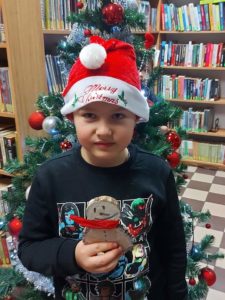 Na zdjęciu: Chłopiec trzyma w ręku bałwanka wykonanego z dwóch plasterków drzewa. Na głowie ma czerwoną czapkę Mikołaja z napisem Merry Christmas. W tle choinka i regały z książkami.