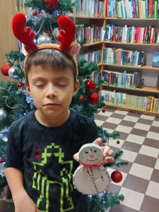 Na zdjęciu: Chłopiec trzyma w ręku białego bałwanka, wykonanego z dwóch plasterków drzewa. Chłopiec ma na głowie opaskę z czerwonymi rogami renifera i zamknięte oczy. W tle choinka i regały z książkami.