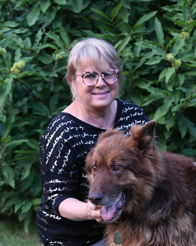 Na zdjęciu, na tle zieleni, Małgorzata w kolorowych okularach i bluzce z połyskującymi napisami. Klęczy obok psa rasy owczarek niemiecki i głaszcze go pod szyją.