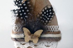 Zdjęcie przedstawia przestrzenny owalny przedmiot przypominający pisankę. Drewniane jajko owinięte jest w połowie koronkami i kolorową włóczką, która utrzymuje czarne nakrapiane piórka. Na środku pisanki przymocowany jest, za pomocą zwykłego sznurka, niewielki drewniany motyl.