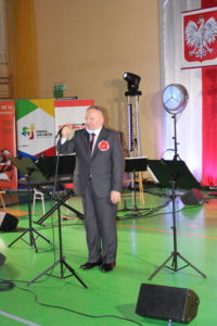 Na zdjęciu: Mężczyzna w garniturze z biało-czerwonym kotylionem przemawiający do mikrofonu.