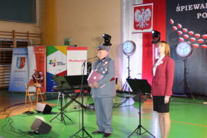 Na zdjęciu: Mężczyzna w wojskowym stroju stoi przy statywie z mikrofonem oraz kobieta w czerwonej marynarce i czarnej spódnicy.