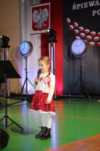 Na zdjęciu: Mała dziewczynka z warkoczykami, ubrana w białą bluzkę i białe rajstopy oraz czerwoną kwiecistą spódniczkę. W ręku trzyma mikrofon. W tle godło Polski.