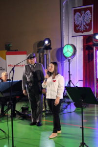 Na zdjęciu: Dziewczynka w białej bluzce i czarnych spodniach śpiewa do mikrofonu. Obok stoi mężczyzna w wojskowym mudurze. W tle godło Polski.