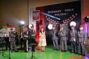 Na zdjęciu: Siedmiu mężczyzn w wojskowych mundurach śpiewają, pośród nich stoi kobieta w jasnej sukience. W tle Patriotyczna dekoracja z napisem: Śpiewamy Tobie Polsko!