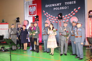 Na zdjęciu: Mężczyźni w wojskowych mundurach oraz dwie kobiety. W ręku ich są mikrofony - śpiewają. W tle patriotyczna dekoracja z napisem Śpiewamy Tobie Polsko!