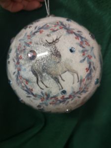 Na zdjęciu: Biała kula - bombka, ozdobiona dookoła niebieskoczerwonym roślinnym motywem. W śwrodku dwa jelenie.