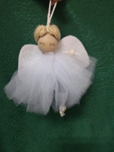 Na zdjęciu: Na ciemnozielonym tle aniołek z główką z drewnianej kulki. Aniołek ma narysowane zamknięte powieki, białe filcowe skrzydełka i białą sukienkę.