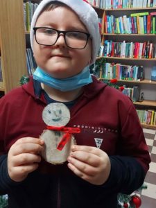 Na zdjęciu: Chłopiec w okularach i bordowej bluzie trzyma w ręku bałwanka wykonanego z dwóch plasterków drzewa. Na głowie ma czerwoną czapkę Mikołaja. W tle choinka i regały z książkami.