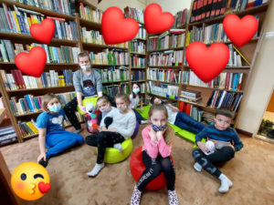 Na zdjęciu: Dzieci siedzą na kolorowych siedziskach, na podłodze. Są radosne. Zdjęcie ozdabiają czerwone serca.