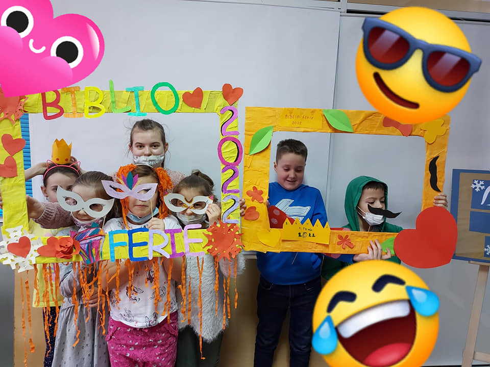 Na zdjęciu: Dzieci trzymają dwie kolorowe ramy. Jedna z nich ma napis BIBLIO FERIE 2022. Dzieci mają na oczach maski i są bardzo radosne. Zdjęcie zdobią kolorowe emotikony.