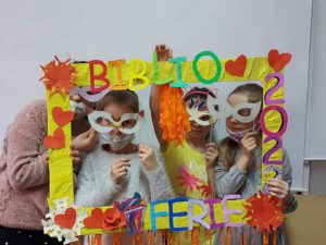 Na zdjęciu: W żółtej, ozdobnej ramie z napisem BIBLIO FERIE 2022 są cztery dziewczynki. Jedna dziewczynka w ręku trzyma pomarańczowe paski bibuły. Wszystkie mają maski na oczach.