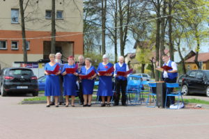 Na zdjęciu: Kobiety i mężczyżni, ubrani na niebiesko trzymają w rękach bordowe okładki. Śpiewają.