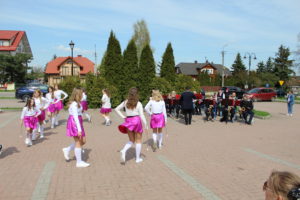 Na zdjęciu: Dziewczynkiw białych bluzkach, różowych spódnicach i białych długich skarpetkachwykonują układ taneczny z jedną ręką opartą o biodro, w drugiej trzymają pałeczki.