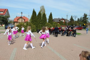 Na zdjęciu: Dziewczynki w białych bluzkach i różowych spódnicach, mają w rękach pałęczki. W tle orkiestra dęta.
