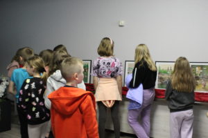 Na zdjęciu: Dzieci stojące tyłem do obiektywu, oglądają kolorowe ilustracje w czarnych ramach
