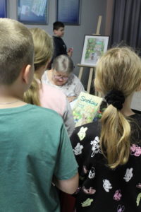 Na zdjęciu: Kilkoro dzieci stojących tyłem do obiektywu oraz kobieta w okularach pisząca w książce.