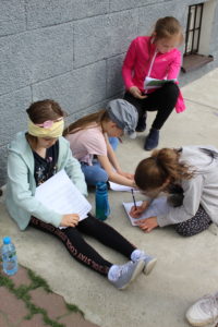 Na zdjęciu: Cztery dziewczynki siedzą na chodniku i piszą na kartkach.