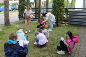 Na zdjęciu: Dzieci siedzą na trawie i piszą na kartkcha.