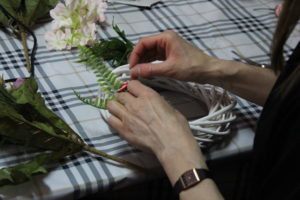 Na zdjęciu: Ręce kobiety, która układa listki na białym wiklinowym wianku.