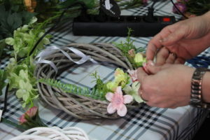 Na zdjęciu: Ręce kobiety, która układa listki i kwiatki na szarym wiklinowym wianku.