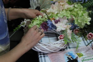 Na zdjęciu: Ręce kobiety, która układa kwiatki na białym wiklinowym wianku.