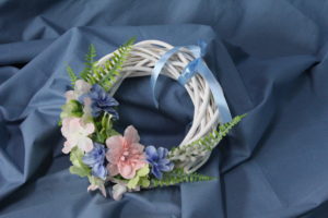 Na zdjęciu: Wianek z kolorowych, sztucznych kwiatów z niebieską wstążką na niebieskim tle.