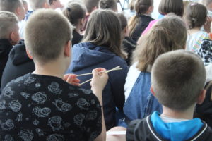Na zdjęciu: Grupa dzieci siedzących tyłem do obiektywu, trzymająchych w rękach pałeczki do jedzenia.