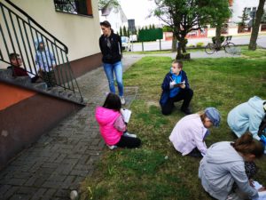 Na zdjęciu: Dzieci siedza na trawie i piszą na kartkach. Kobieta w czarnej bluzie stoi i z nimi rozmawia.