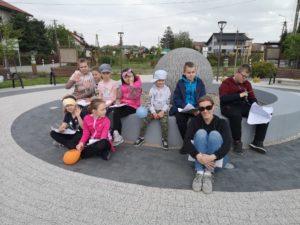 Na zdjęciu: Grupa zadowolonych dzieci, wraz z kobietą w ciemnych okularach siedzą przy fontannie w kształcie kuli.