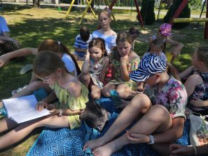 Dzieci siedzą na kocu i słuchają dziewczynki, która czyta książkę