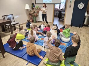 Dzieci siedzą na dywanie i słuchają prowadzącej zajęcia.