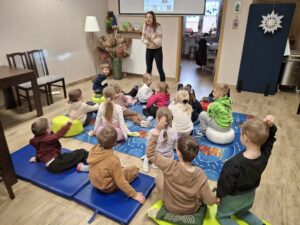 Dzieci siedzą na dywanie i słuchają prowadzącej zajęcia.