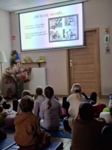 Dzieci siedzą na dywanie i oglądają prezentację multimedialną.