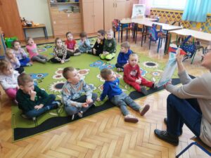Dzieci siedzą na podłodze i oglądają ilustracje w książce, którą pokazuje kobieta.