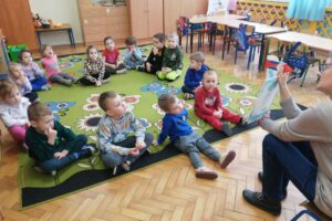 Dzieci siedzą na podłodze i oglądają ilustracje w książce, którą pokazuje kobieta.