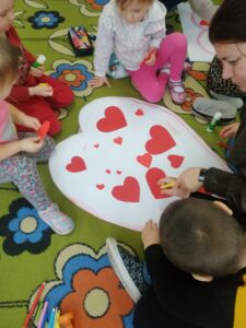 Dzieci siedzą na podłodze i przyklejają czerwone serduszka do konturu serca.
