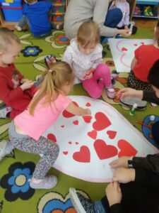 Dzieci siedzą na podłodze i przyklejają czerwone serduszka do konturu serca.