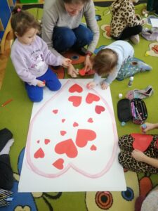 Dzieci siedzą na podłodze i przyklejają czerwone serduszka do konturu serca.