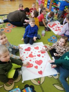 Dzieci siedzą na podłodze i przyklejają czerwone serduszka do konturu serca.