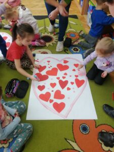 Dzieci siedzą na podłodze i przyklejają czerwone serduszka do konturu serca.