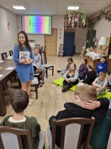 Grupa siedzących dzieci oraz stojąca kobieta z ksiązką w reku.