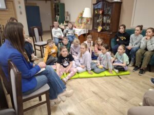 Kobieta oraz grupa dzieci siedzących na sofach, krzesłach i podłodze.