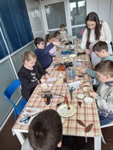 Grupa dzieci i instruktorka, podczas warsztatów rękodzieła- zające.