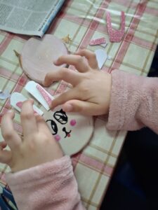 Dziecięce dłonie przyklejają uszy zajączkowi, wykonanemu z plastra drzewa.