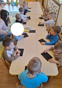 Dzieci siedzące przy stoliku, wydrapują patyczkami w czarnych kartkach.